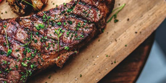 A grilled steak garnished with herbs sits on a wooden cutting board next to roasted garlic. - Matrix Sciences